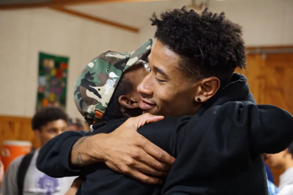 Two black men with medium-deep are shown hugging one wearing a camoflauge hat and the other has short coily hair.