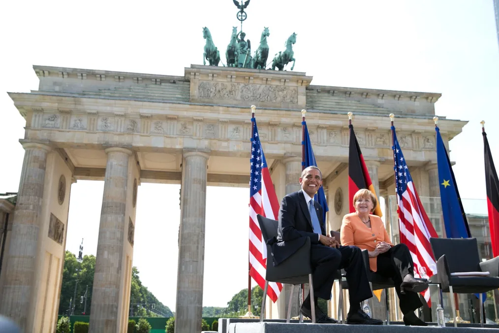 President Barack Obama and Angela Merkel sit in black chairs with flags representing different countries behind them. There is also a large monument with columns and green horses in the background.