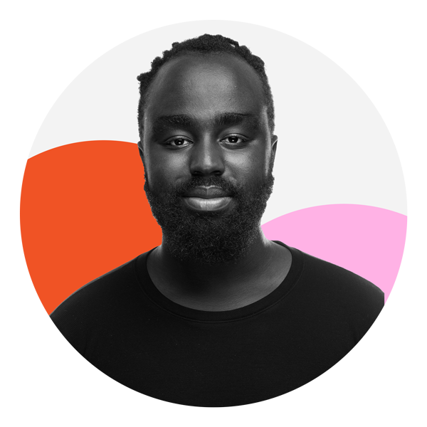 A deep skin toned Black man is smiling with his mouth closed. He is wearing a dark t-shirt. He has short Twisted hair and a full mustache and beard. The photo is black and white and the background features two colored circles, one which is orange and the other pink .