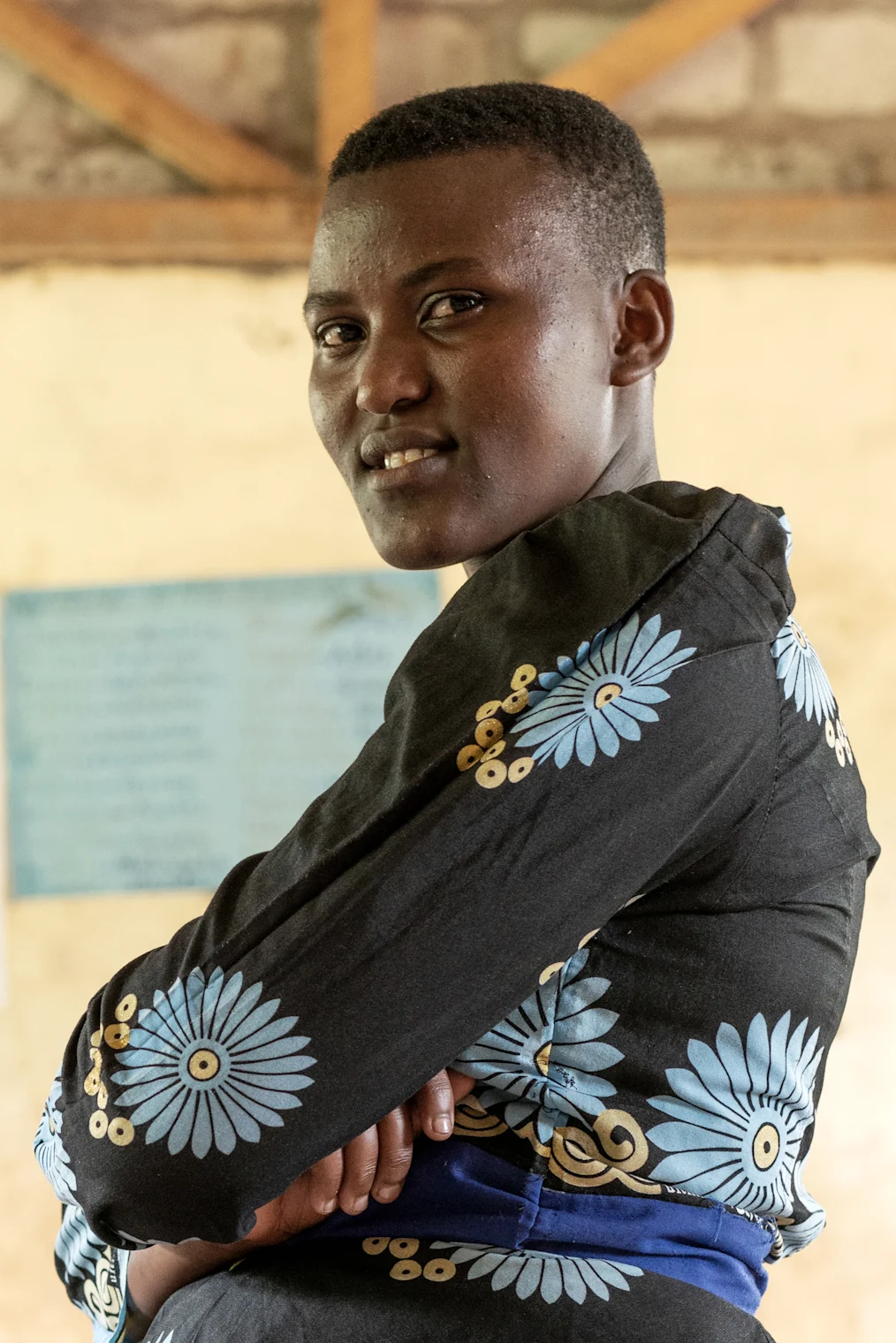 Judith Godfrey, a young Tanzanian woman with a dark skin tone, poses in a classroom with her arms crossed. She wears a black dress with blue flowers and has short hair.