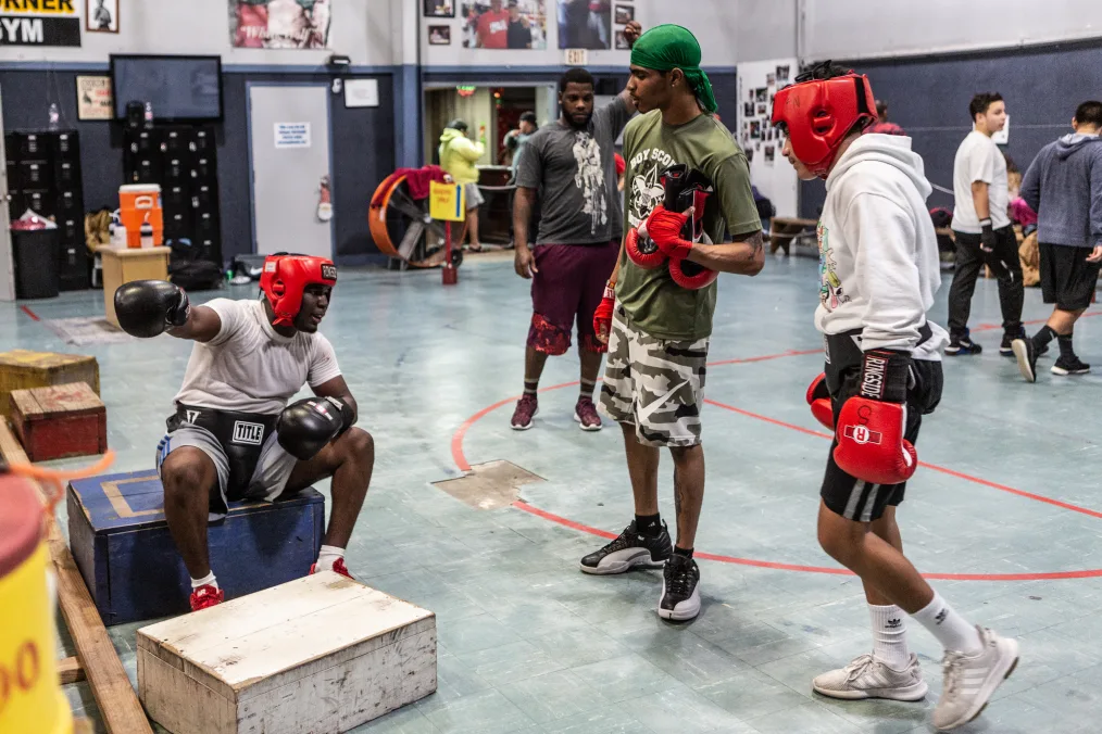 3 young men, the main subjects of the photo, have a conversation. The setting is a boxing gym. They are of medium to deep skin tones and casual athletic attire on mixed with boxing gear.   