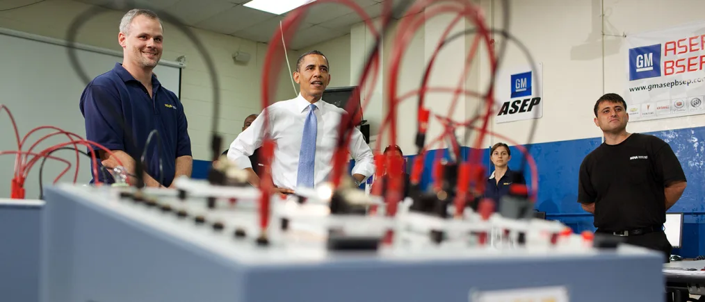 President Obama stands with a group of people with various skin tone. The camera captures a red and black wired switchboard out of focus.