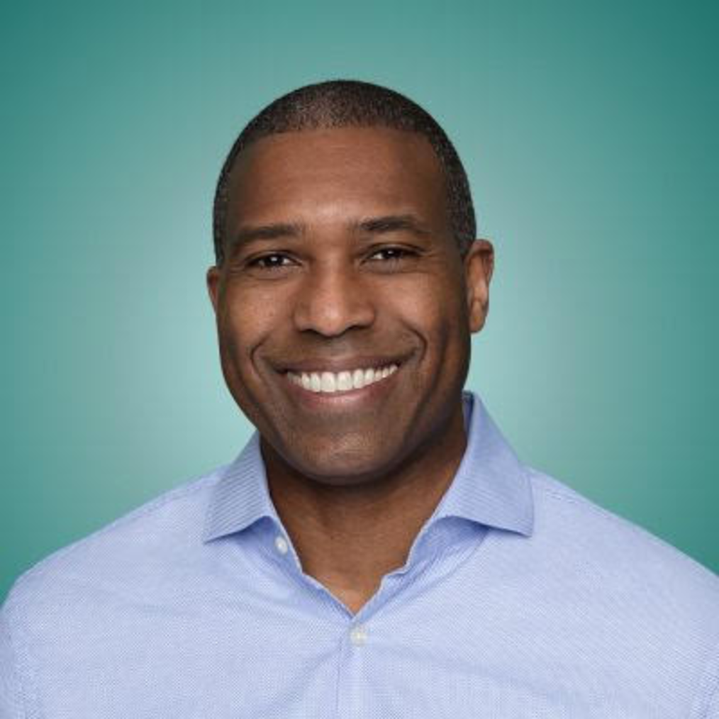 A Black man with short hair smiles broadly. He wears a blue button-down shirt and there is a teal background behind him.