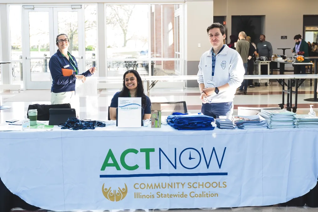 Scholar Nikki Gillan, a woman with a medium skin tone and dark brown hair sits behind a table that reads, “Action Now” and “Community Schools Illinois Statewide Coalition.” Next to her is a man with a light skin tone and blonde hair. Behind her is a woman with light skin tone, brown hair, and glasses. 