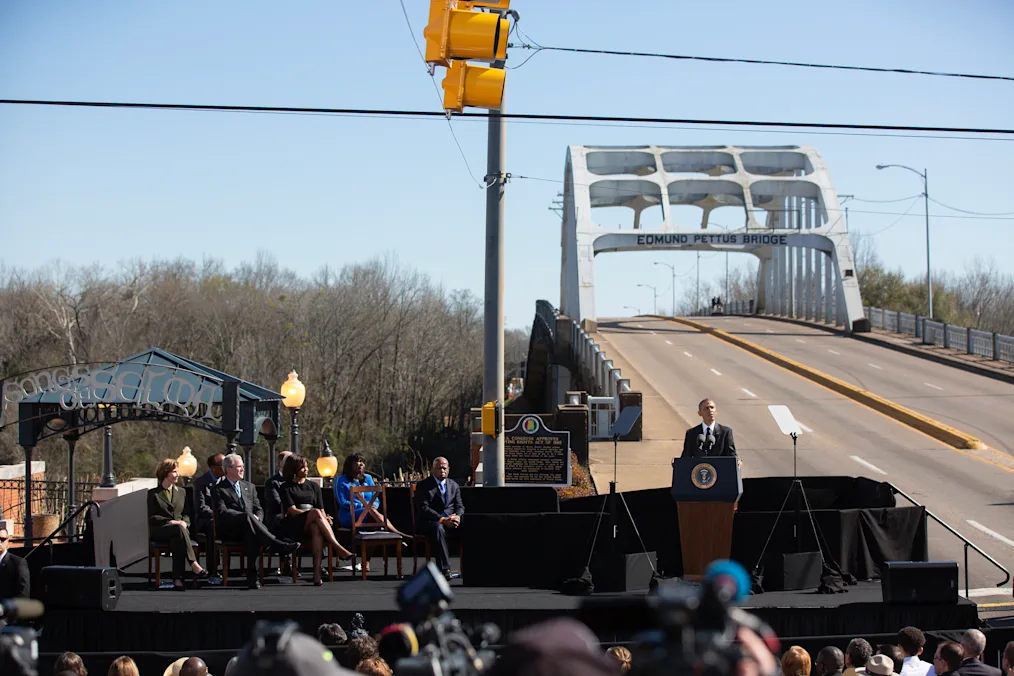 An image of President Barack Obama giving a speech at the 50th Anniversary of the Selma marches in Selma, AL. President Obama is standing in front of Edmund Pettus Bridge. It is daytime. Next to President Obama is a seating area where the First Lady Michelle Obama, President George W. Bush, and First Lady Laura Bush, and Congressman John Lewis are seated.