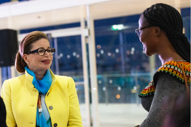 Valerie Jarrett smiles as she stands next to Beverly Ndifoin Niyang, a Black woman with a deep skin tone. She is wearing glasses and braids. Valerie Jarrett is wearing a yellow blazer with a blue scarf.