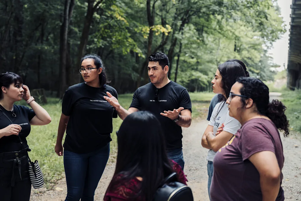 José Patiño, a man with a medium skin tone, stands in the middle of a group of young people in August 2019 talking about their experience in Selma, Alabama, learning about the civil rights march there. All have medium skin tones and are standing outdoors. He and the woman on his left are wearing a shirt that reads, “the march continues.”