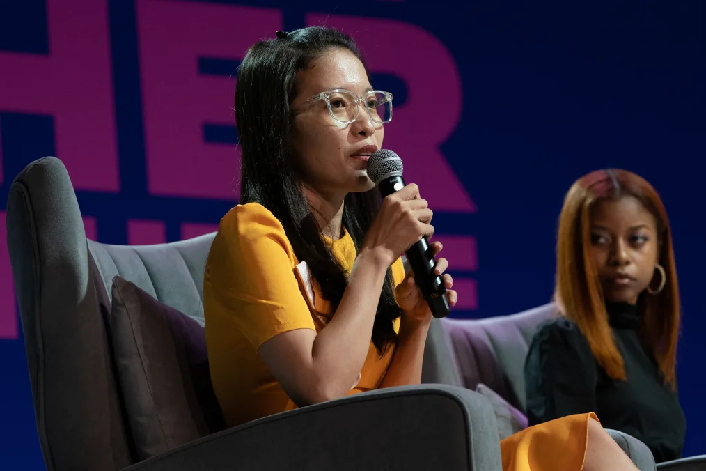 Kim Nho Nguyen, a light skin woman with dark hair, holds a microphone close to her mouth as she speaks at the Girls Opportunity Alliance Get Her There campaign event. She is wearing a yellow dress and clear glasses. On the right, Addison Belhomme listens attentively.