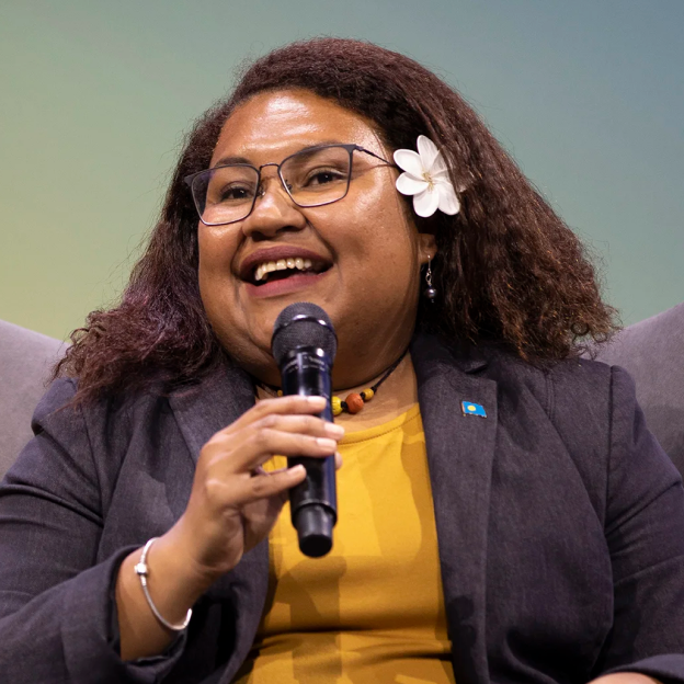 A young woman with a medium-deep skin tone sits and smiles while speaking into a microphone. She wears a yellow top and a gray blazer, glasses and a flower in her hair. In the background, a blue, green and yellow gradient backdrop. 