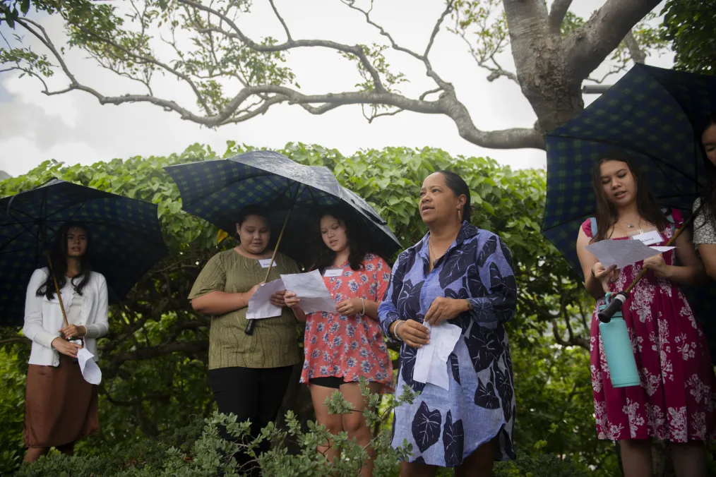 A group of young women led by a older woman who all have what could be island, latin, or asian facial features. They are standing in an enviorment surrounded my small trees and bushes. Some are holding umbrellas or papers. Most of them are wearing dresses with a tropical flower or plant on them.  