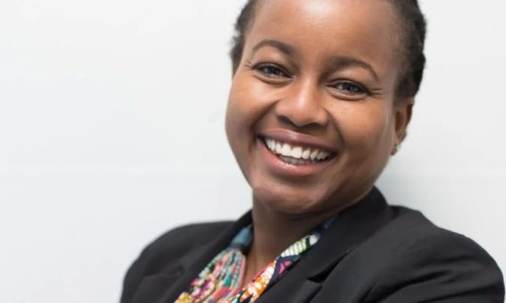 Wambui Kinya, a black woman with a medium-deep skin tone wearing a black suit jacket and colorful patterned shirt, smiles toward the camera.