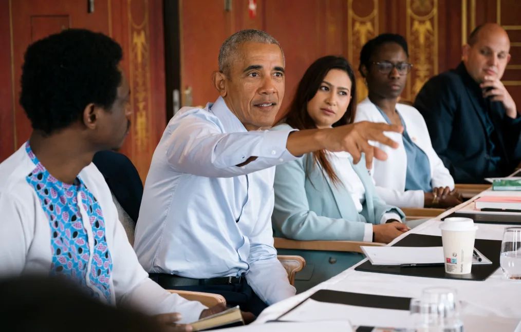 President Obama sits at a table between two men and women with a range of skin tones and clothing styles. The President’s right arm is extended as he looks away from the camera. 