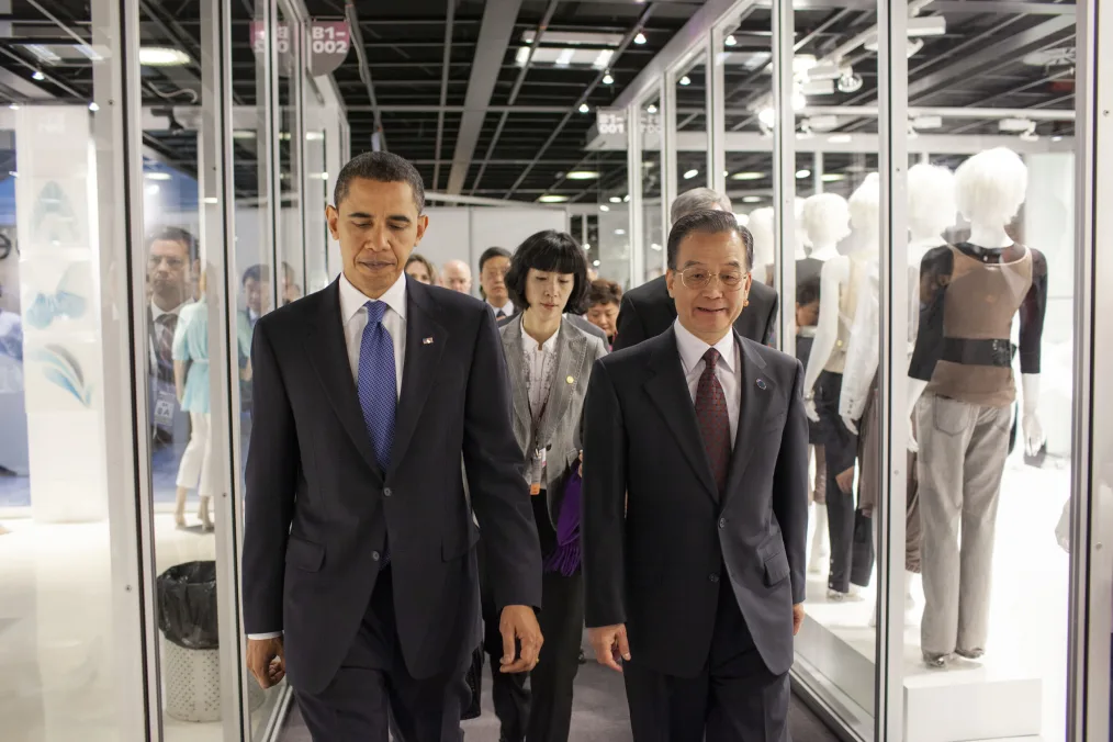 President walks down a hallway with clear cases filled with maniquens with a group of older individuals with various skin tones in buisness attire  