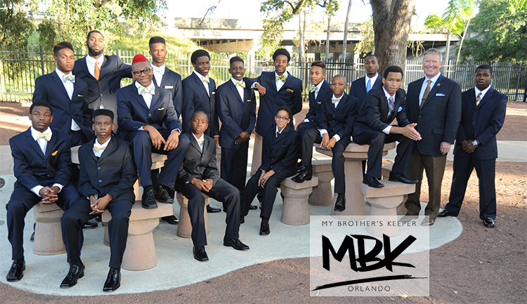 A group of men, most young-looking and Black, wear suits and pose for the camera in an outdoor area. A graphic in the lower right says "My Brother's Keeper, MBK, Orlando." 