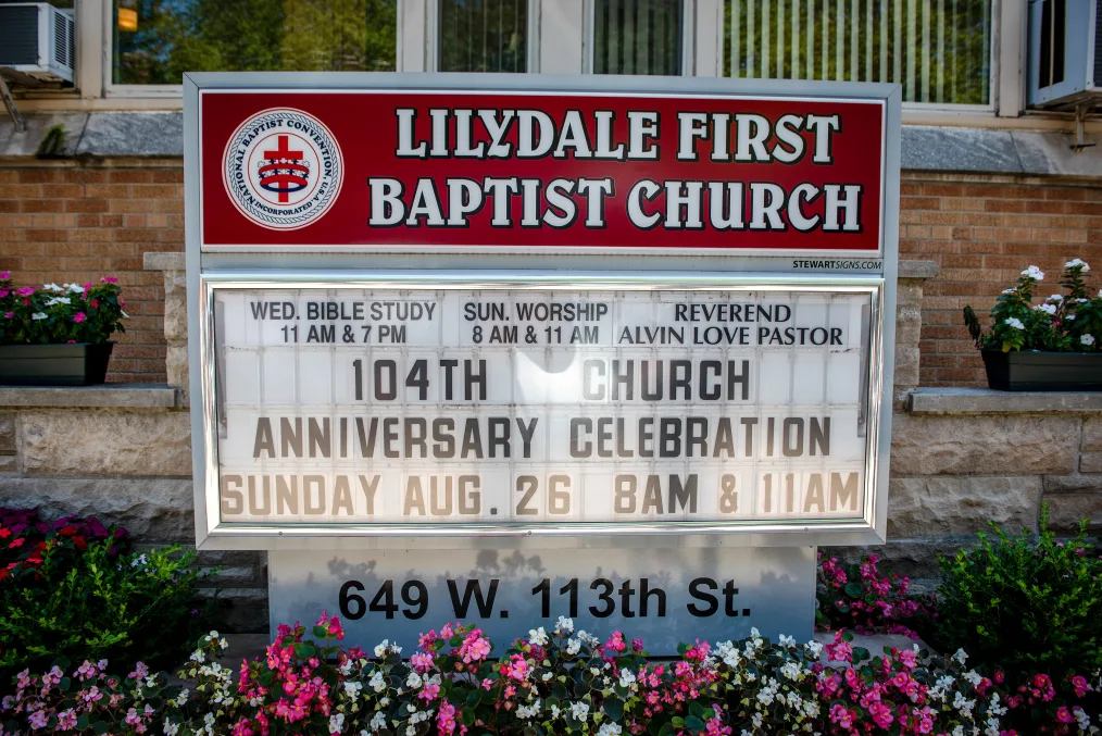 A red and white Lilydale First Baptist Church sign. It has important event times underneath such as bible study. The base of the sign reads "649 W. 113th St.