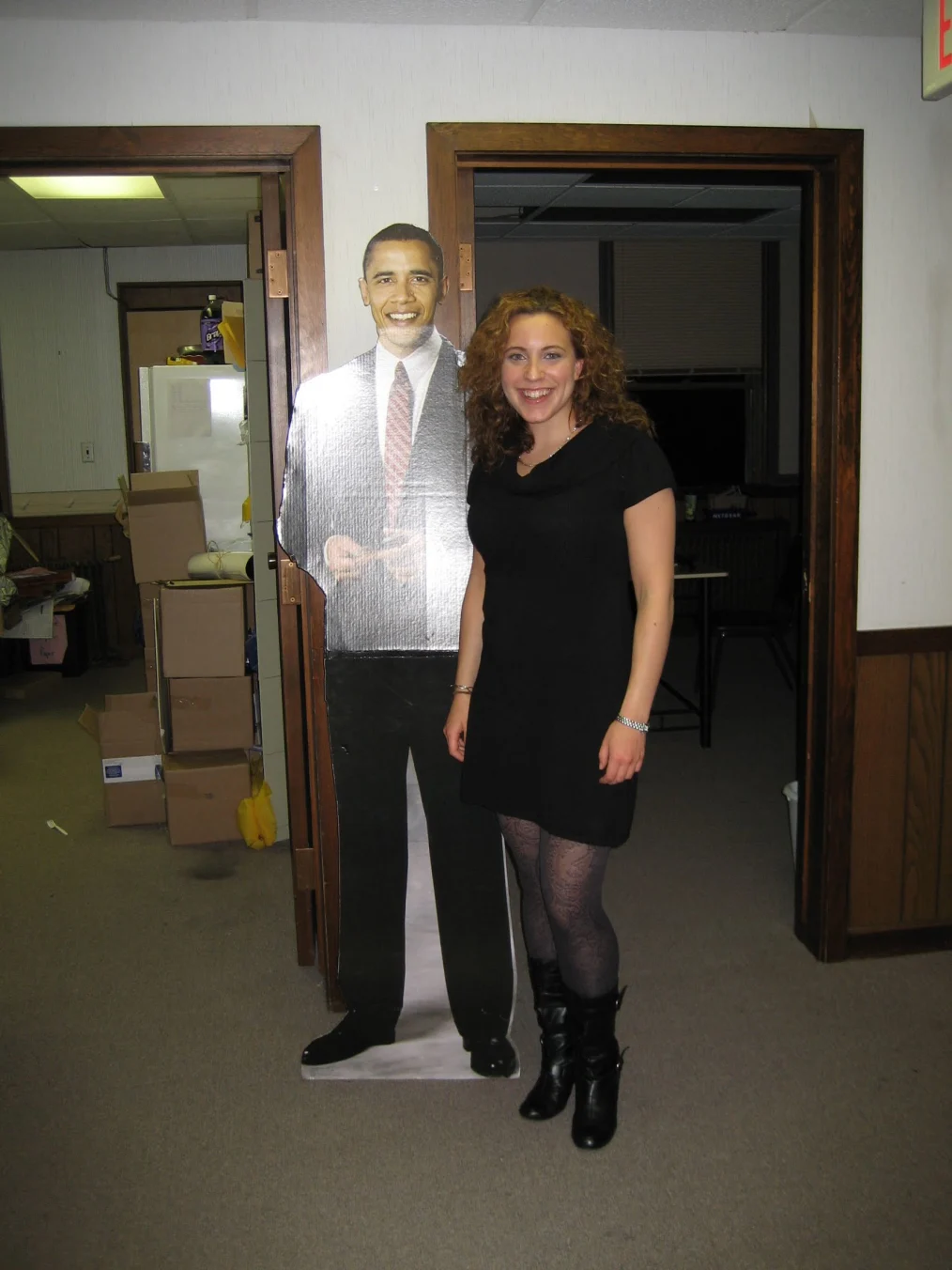 Beth Pollack is standing in an Obama campaign office smiling at the camera. Beth has very curly brown hair with gold highlights. She is wearing a black dress that hits above her knees, black lace tights, and black boots, with buckles, that end at her lower calf.  On both of her wrists are singular silver bracelets. Next to Beth is a life size cardboard cutout of Barack Obama. The cutout is an image of Barack Obama from the year 2007. In the cardboard cutout Barack Obama is wearing a black suit with red tie, black shoes, and is smiling at the camera. In the campaign office, there are two open wooden doors revealing two rooms. Inside the room on the right is darkness because the lights are off. Inside the room on the left are stacked boxes, a chair covered in coats, a refrigerator with a purple drink sitting on top of it, and a lone fork on the floor.