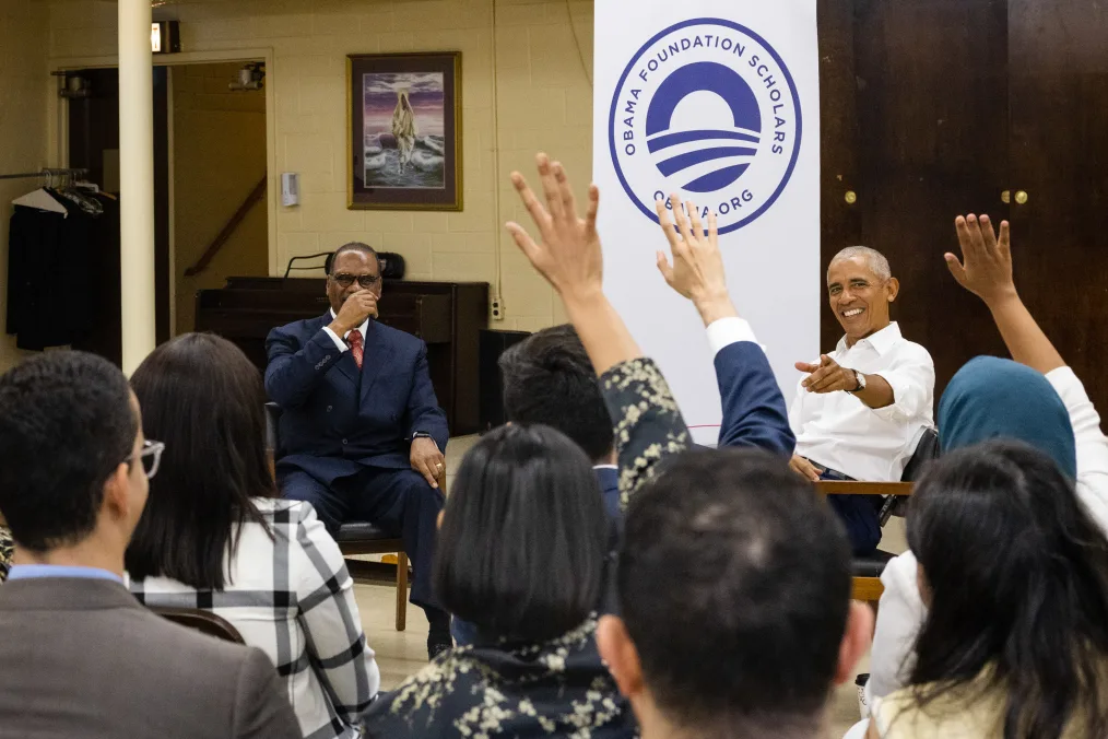 President Obama sits next to Pastor Alvin Love, a Black man with a deep skin tone, as he points at several 2023 Obama Scholars. The scholars' backs are to the camera. Behind President Obama is a sign that reads, “Obama Foundation Scholars” and “Obama.org.” 