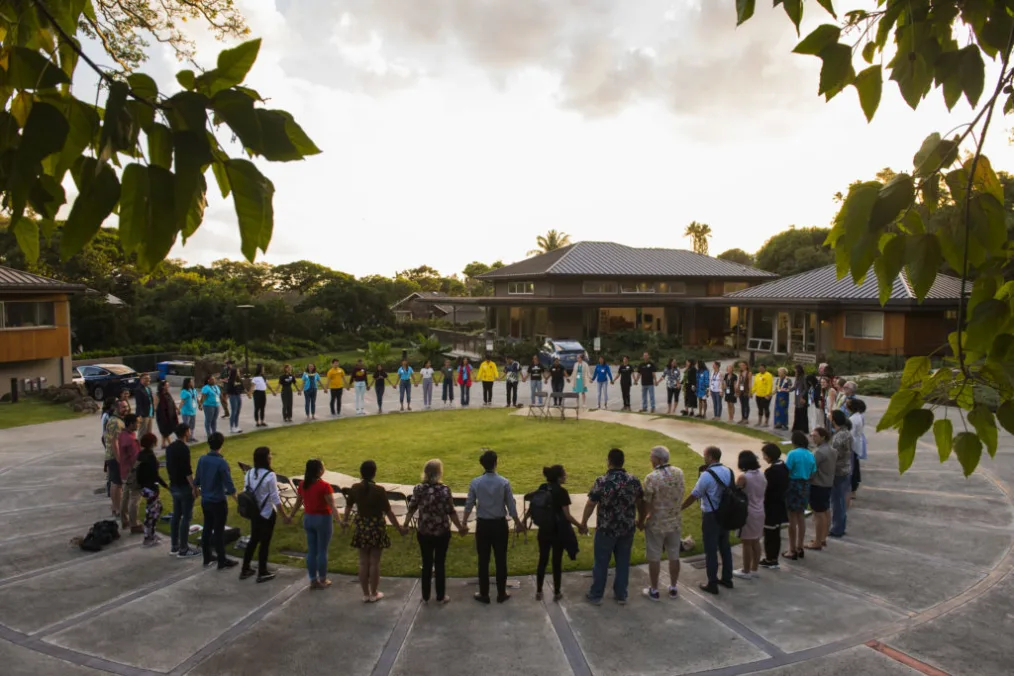 On a sunny day, a group of people with a range of light to medium deep skin tones hold hands and create a wide circle on the outskirts of a green grass patch. 