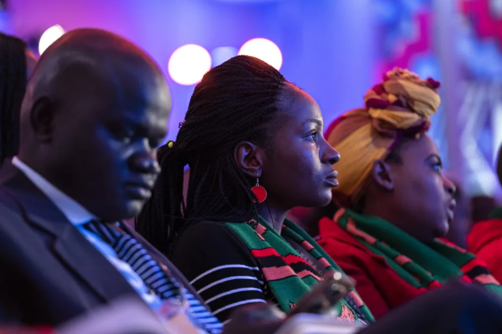 The camera foucses on a man with a deep skin tone dressed in buisness attire and 2 women with deep skin tones wearing african clothing pieces as they look into the distance 