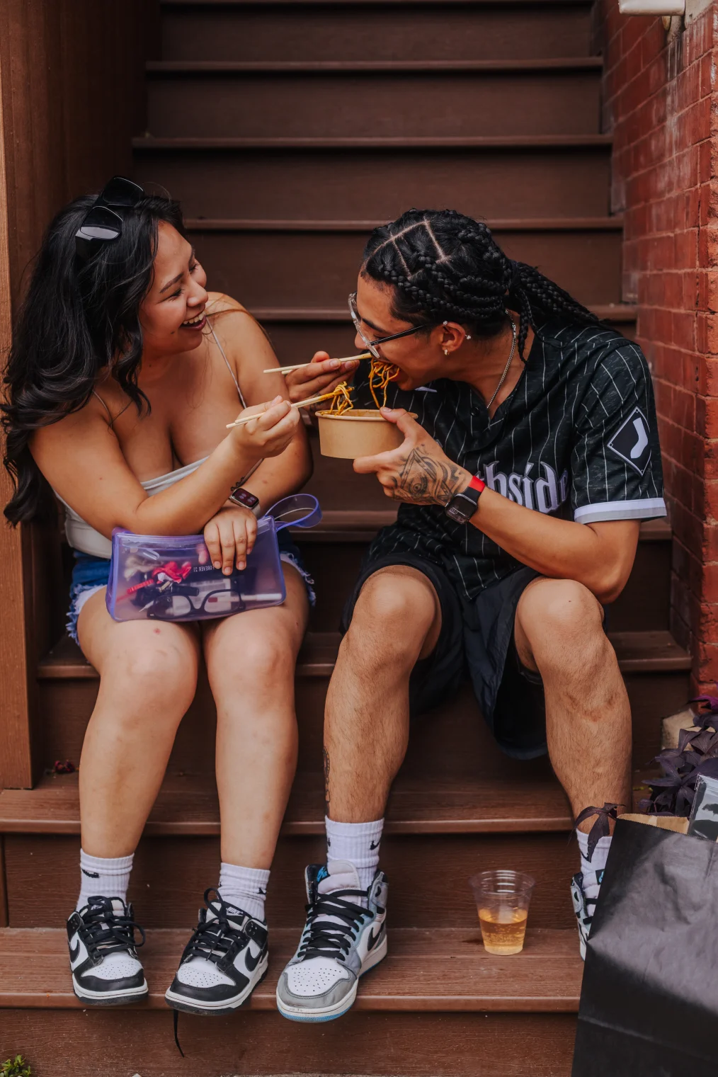 The image is of two people sitting on the steps of an building, laughing and eating a bowl of noodles. The first person is a young woman: she has a light olive complexion and long wavy dark hair with dark sunglasses sitting on top of her head. She is wearing a grey tank top, blue denim shorts, and black and white Nike Air Force sneakers. In her left hand she is holding a purple makeup pouch, in her right hand she is holding a pair of chop sticks over the bowl of noodles. The second person in the picture is a man. He has an olive complexion and long dark braids. He is wearing glasses, a baseball jersey, black track shorts, and grey, white, and black Nike air force sneakers. He is eating from the bowl of noodles with his right hand and holding the bowl of noodles with his left hand. On his left hand is a tattoo and on his left wrist is a red watch. In between his legs, on the steps, is a plastic cup with a beverage inside. To his left, on the steps, are several shopping bags.