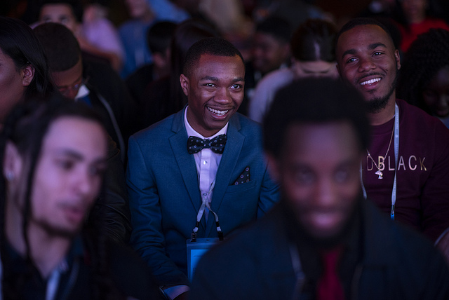 A crowd of men and women who are majorily darker toned and in neat attire at an event. They are seated next to each other in rows. The camera focuses on the middle row where two medium deep toned men share a laugh. 