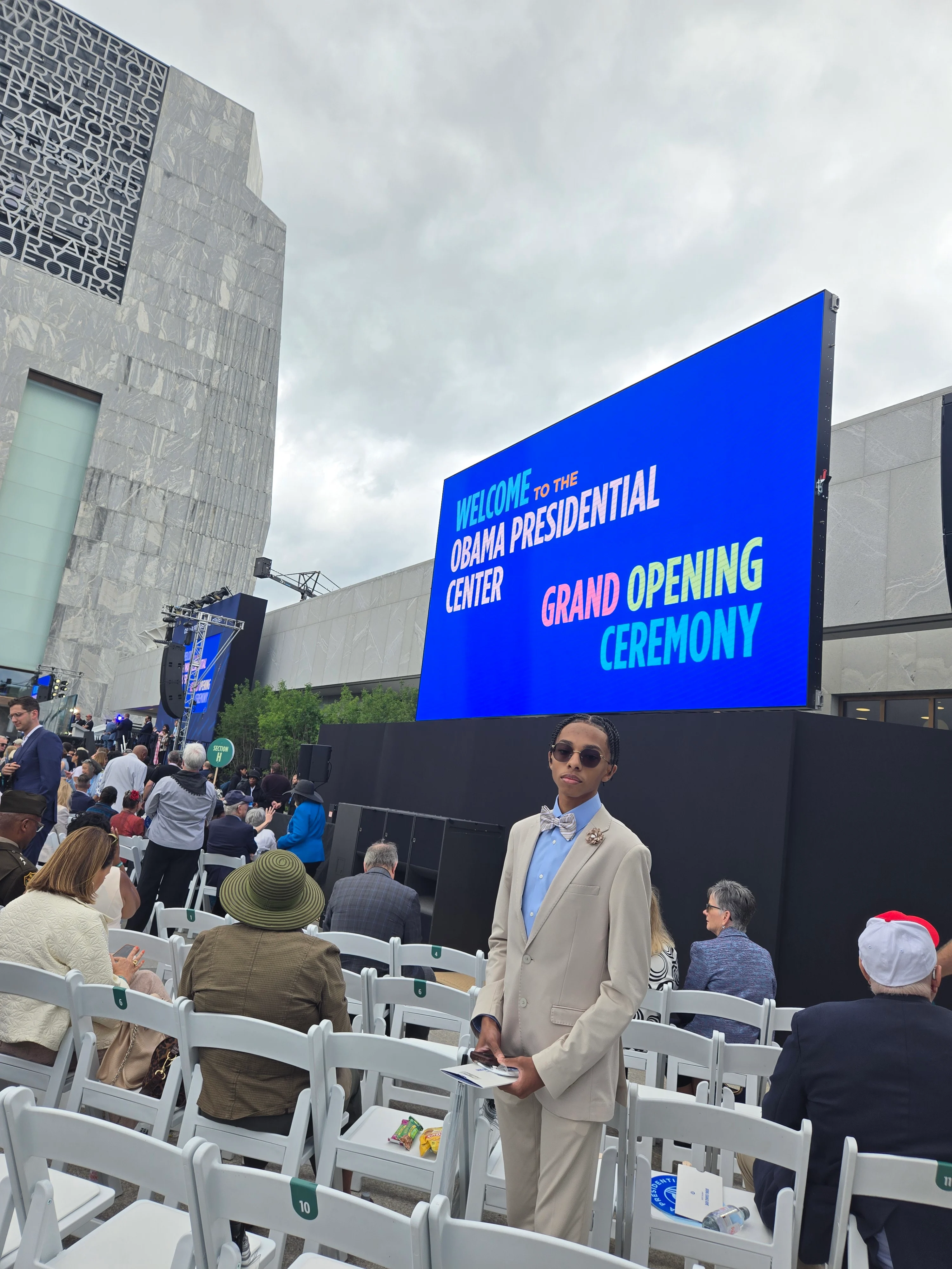 Jonathan Sullen, a 16-year-old Black boy with a light skintone, smiles as he stands in between white chairs at the Obama Presidential Center grand opening ceremony. He is wearing a tan suit and a blue shirt. A cobalt blue monitor in the background reads, “Welcome to the Obama Presidential Center Grand Opening Ceremony.”