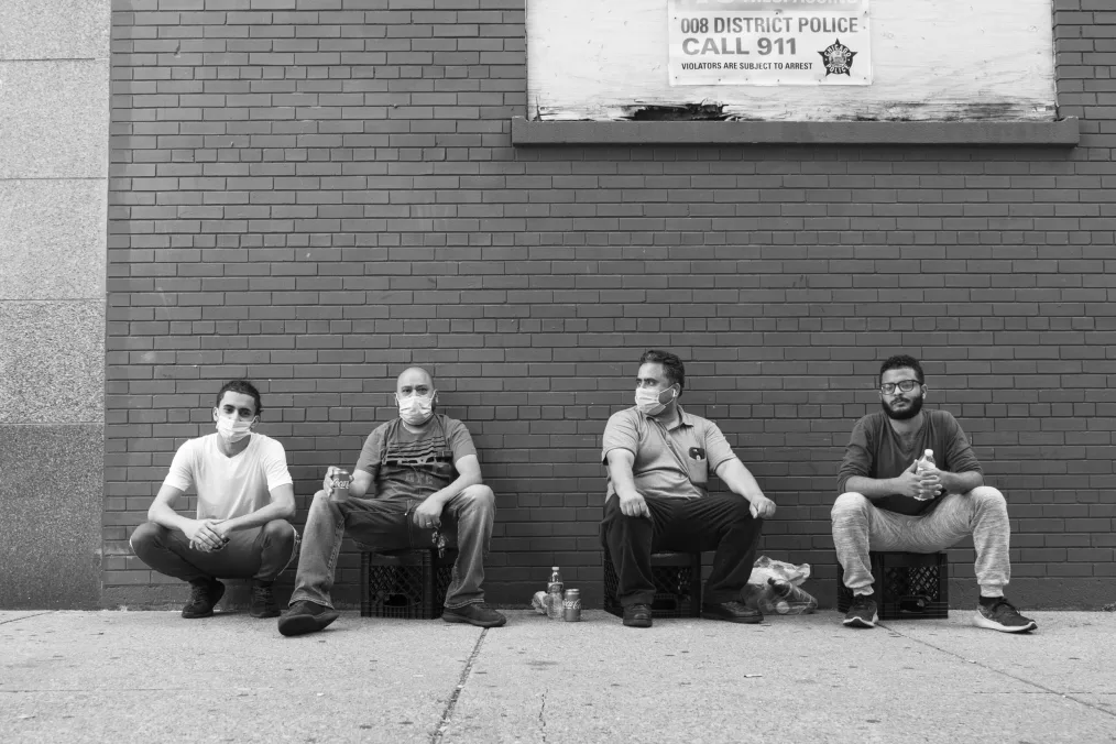 Men sit against brick wall wearing surgical masks. 