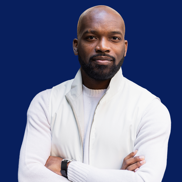 The image is a headshot of Abdul Dosunmu. Abdul has a deep skin complexion, a bald head, brown eyes, and a black goatee. He is wearing a white sweater with a white t-shirt underneath. His arms are folded across his chest. On his left wrist is a black watch. The background of his photo is the color blue.