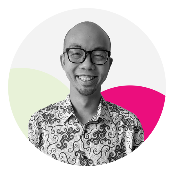 A medium skin toned man with a bald head is facing the camera smiling with his teeth showing. He is wearing a patterned button-up shirt and dark framed glasses. The photo is black and white and the background features two circles, one which is light green and the other dark pink. 