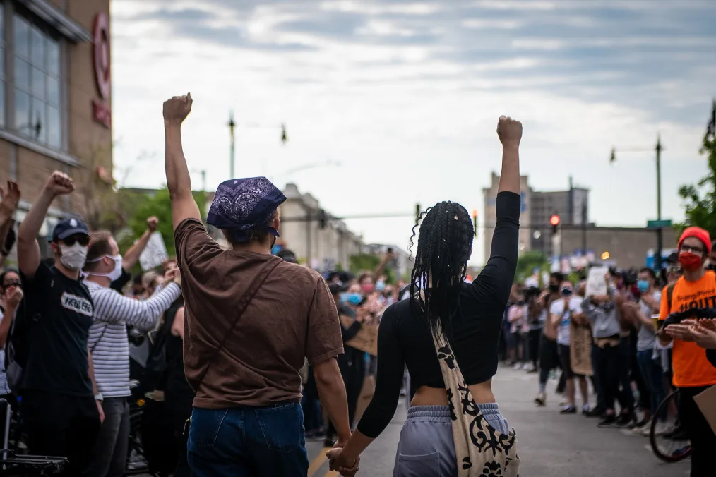 Two people hold hands and their fist in the air. Protesters gather on both sides around them. 