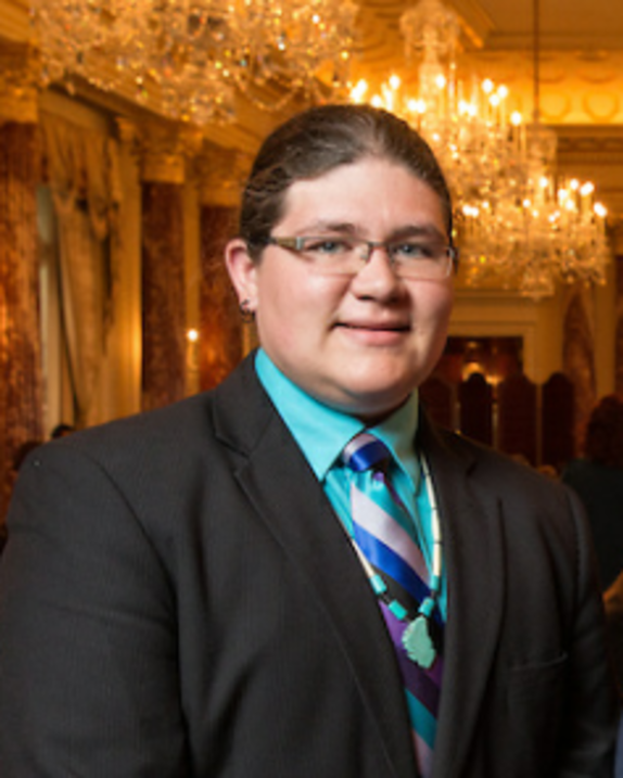 In this picture a man with a light skin tone wearing glasses and a black suit with
a multi-colored tie and a blue shirt underneath poses toward the camera.
