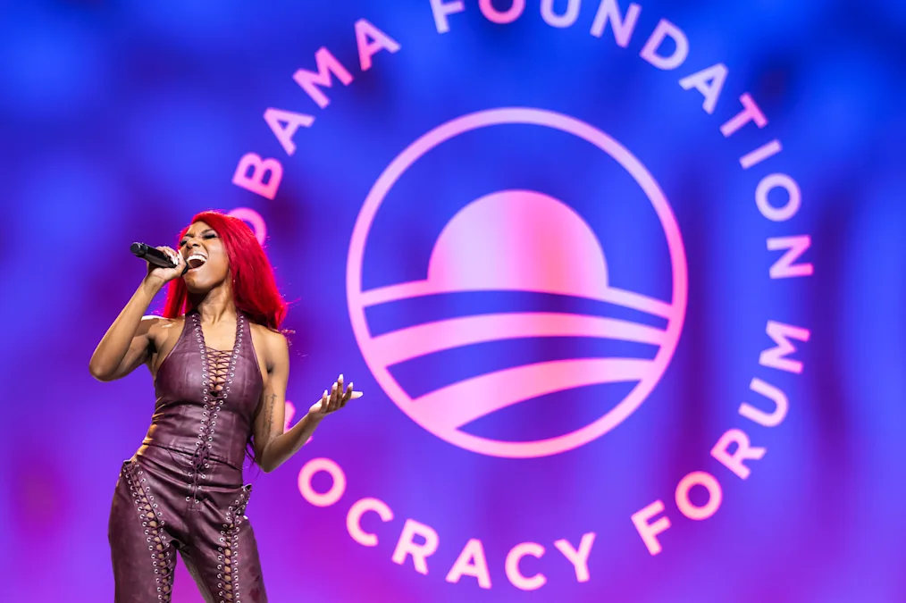 Reyna Roberts, a woman with red hair and a dark skin tone, sings at the 2024 Democracy Forum. She is wearing brown leather. 