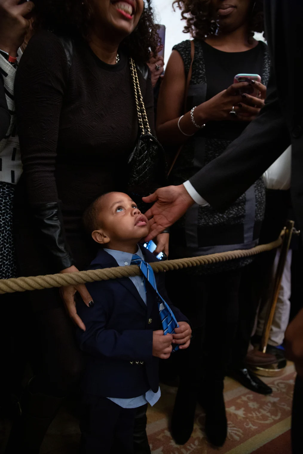 A young boy with a light skin tone looks up as President Obama holds his face. President Obama is out of frame. The boy is wearing a blue suit and tie. People gather behind and watch on. 