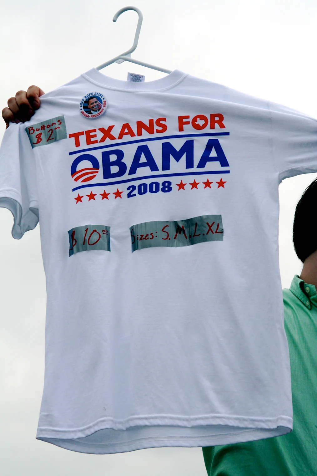 A man with a light skin tone is out of frame as he holds up a white shirt that reads, “Texans for Obama 2008.”