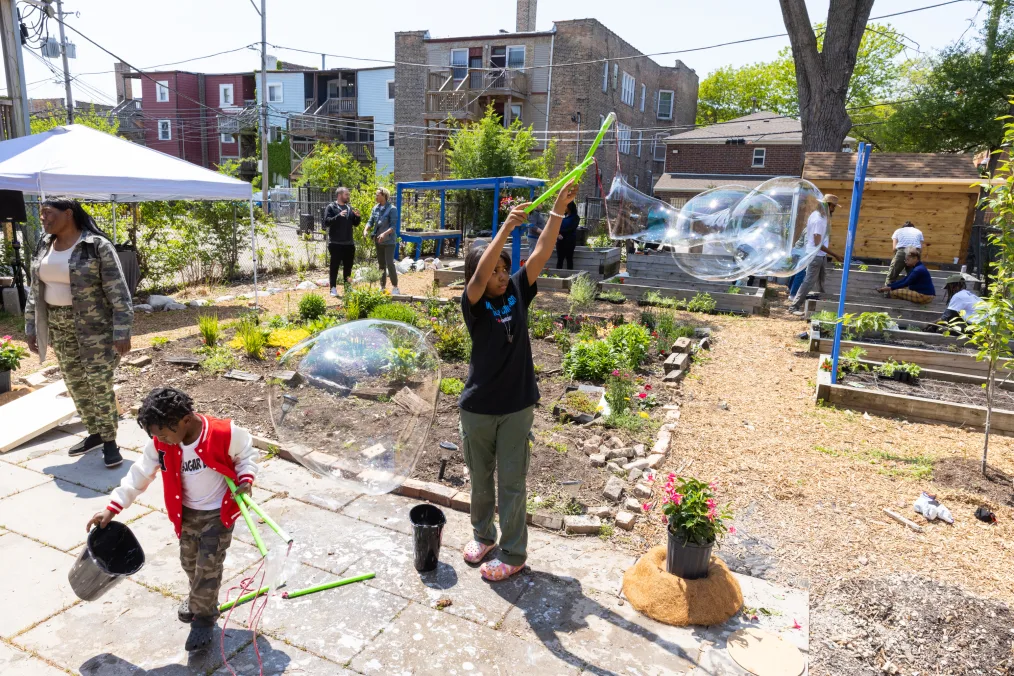 On a sunny day, people with a range of light to deep skin tones gather at a community garden. In the foreground, a  young girl with a deep skin tone plays with bubbles and a young boy with a deep skin tone holds a bucket and jump rope. In the background are several flower beds and people working in the garden. 