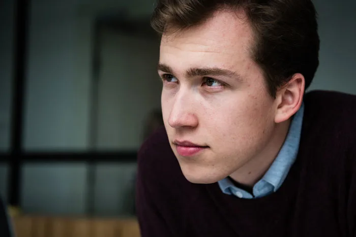 A close-up of a White man looking intently to the left side. He has brown hair and wears a button-down collared shirt under a sweater.