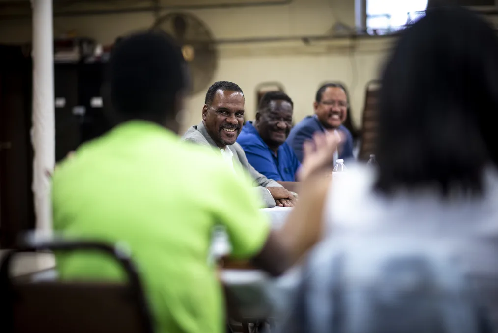 A man with a dark skin tone smiles and sits at a table to talk with a group of people, including Michael Strautmanis in the background.