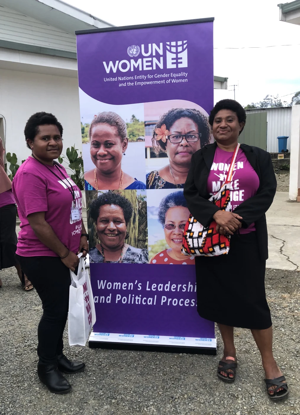 Emma Minimbi, a woman with a medium deep skin tone, holds a closed lip smile as she stands to the left of a UN Women sign. Posing on the right is a woman with a medium deep skin tone with a closed lip smile. Both women have short hair and are wearing purple shirts that read, “Women make change.” The UN Women signage reads, “UN Women, United Nations Entity for Gender Equity and empowerment of women, and Women’s leadership and political process.”