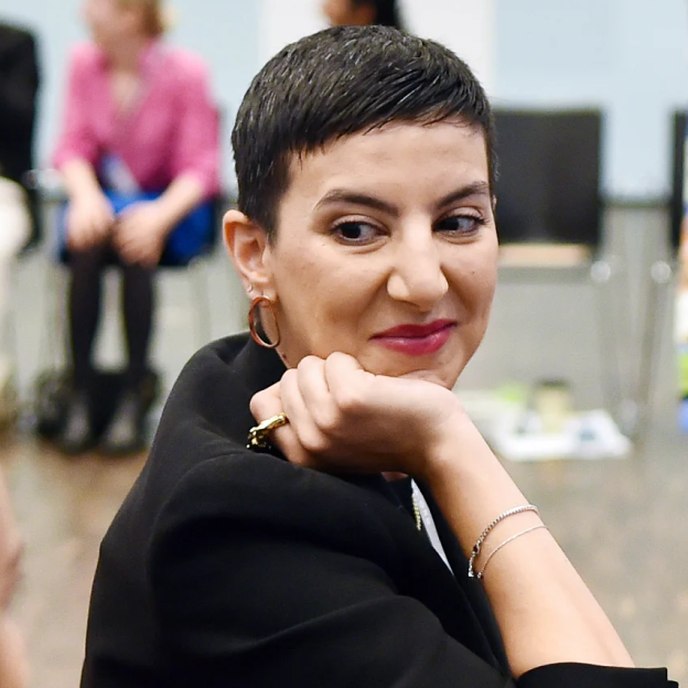 A young woman with a light skin tone and short black hair smiles behind her with her hand on her chin. 