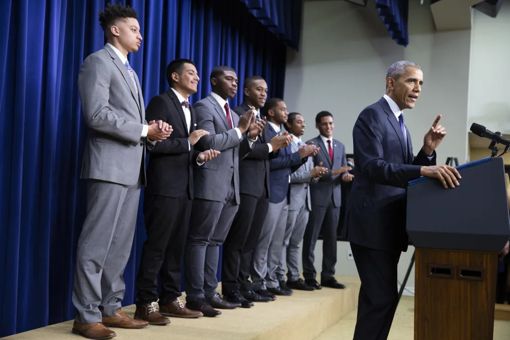 Seven young men with a range of light to deep skin tones stand behind President Obama at a podium. All are wearing a gray, navy, or black suit.