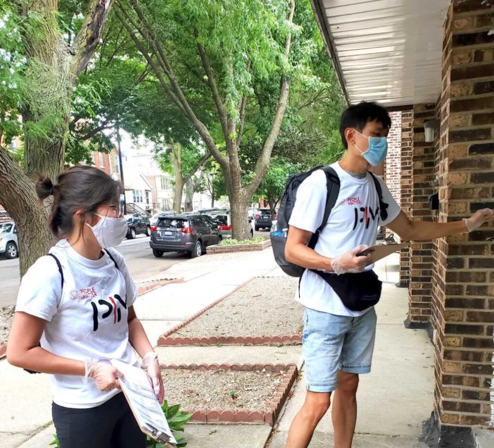 Two young people wearing masks knock on doors to speak with neighbors.
