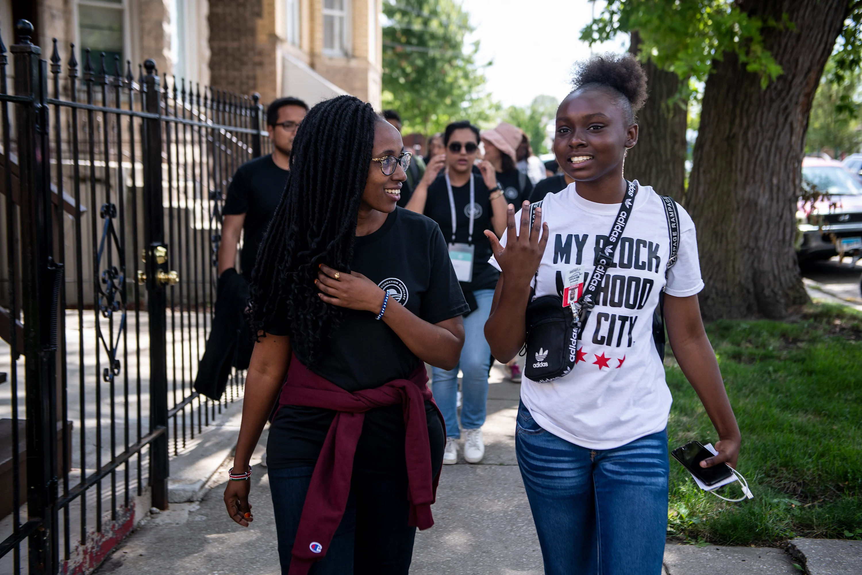 Two women with dark skin tones walk down a street. It’s a sunny day. A group of people with a range of light to dark skin tones walk behind them.