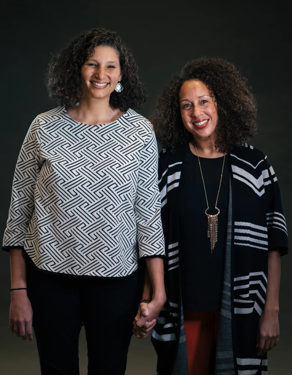 Two women with medium skin tones hold hands and smile toward the camera.