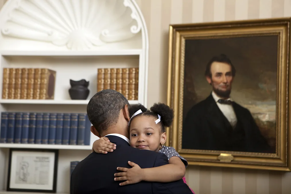 A photo of President Barack Obama holding a little girl in the Oval Office. President Obama’s back is turned to the camera. The little girl is facing the camera. She has a light deep complexion. She has two afropuffs in her hair. They are standing in front of a picture of President Abraham Lincoln.