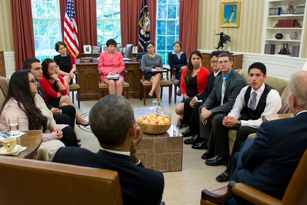 The image is a picture of President Obama speaking to a group of DACA recipients in the Oval Office. President Obama is facing away from the camera. Next to him is former President Joe Biden. Across from him are several White House officials including Valerie Jarrett. Sitting on either side of him are several young people. They are different genders and different races. In the middle of the room is a stone coffee table, on top of the coffee table is a bowl of oranges and several glasses of water. In the background of the photo is a window looking out to the White House Garden. It is sunny outside. The windows are surrounded by deep red curtains. In front of the window is the President's desk and two flags: The American flag and the Flag of the President.