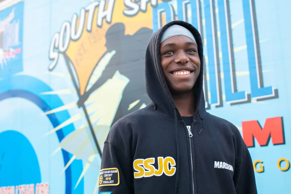 A Black boy with a deep skin tone smiles away from the camera. He is wearing a hoodie that reads, “SSJC” and “Midwest Color Guard Circuit 2018 Finalist.” In the background is a South Shore Drill team mural. 