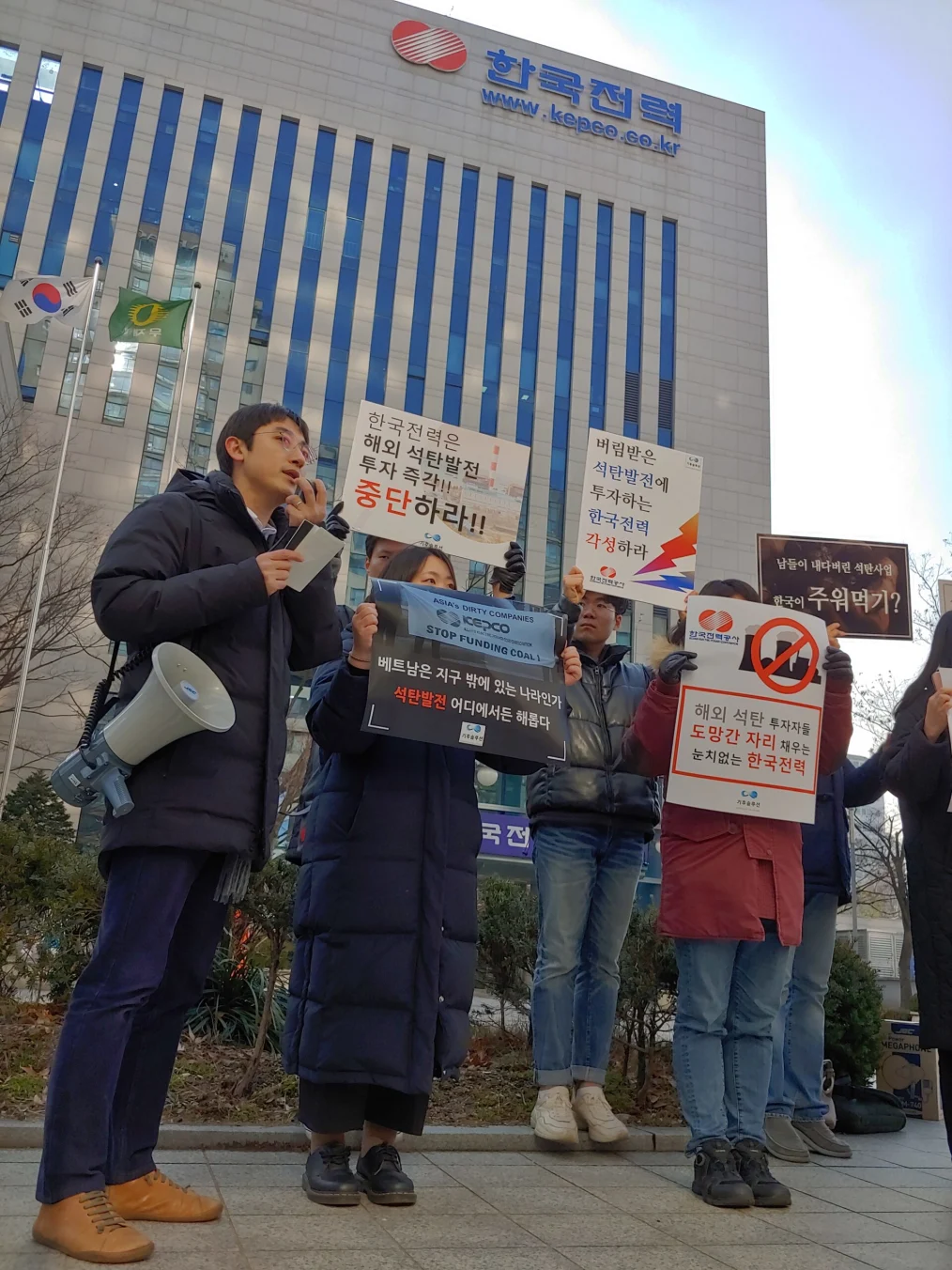 Sejong Youn holds a megaphone as he stands outside next to five other demonstrators at a protest on overseas coal investment. Demonstrators are wearing winter coats and holding signs.