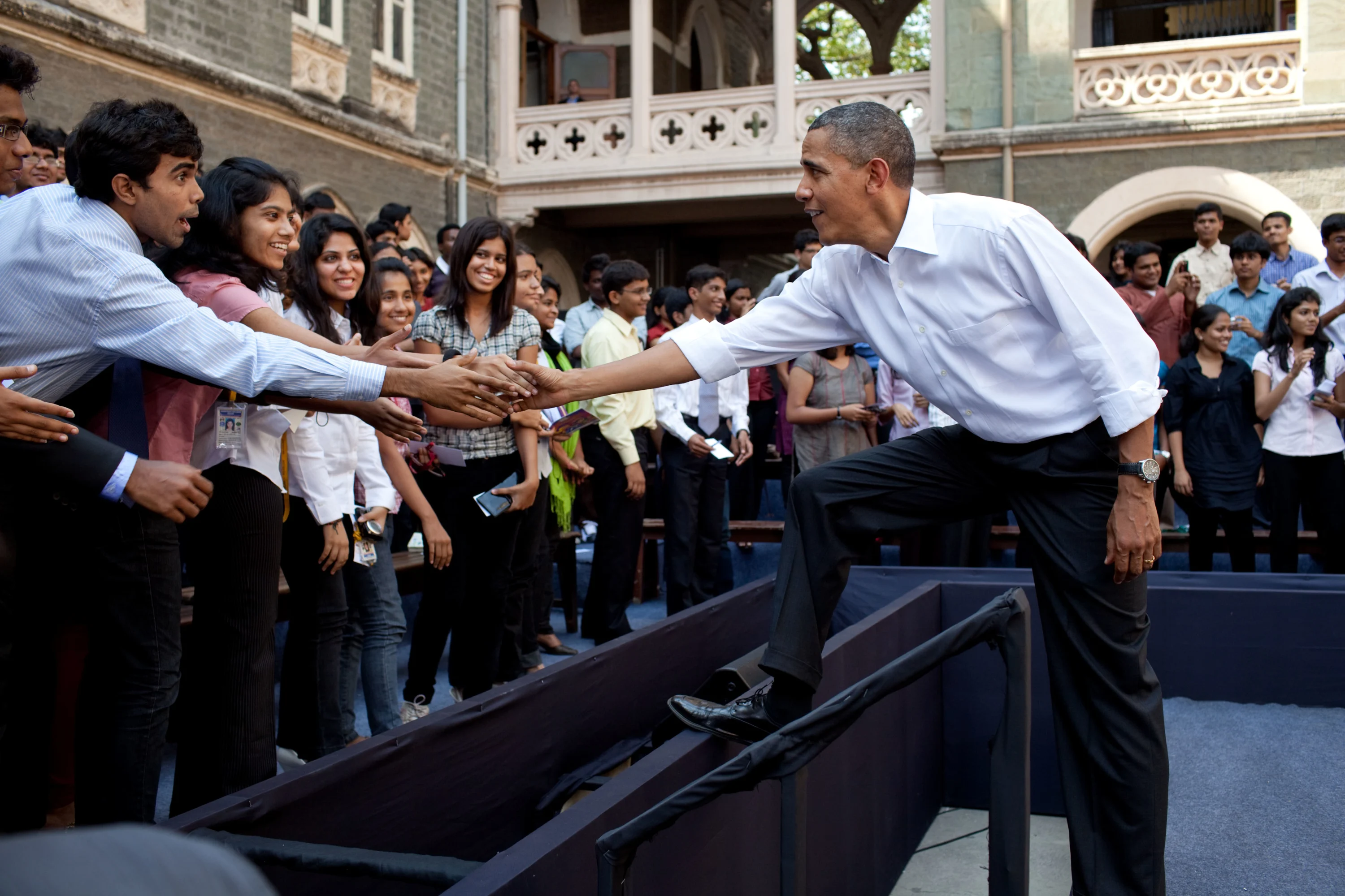 President Obama stands in the middle of a large group of people with various skin tones and shakes hands with them.