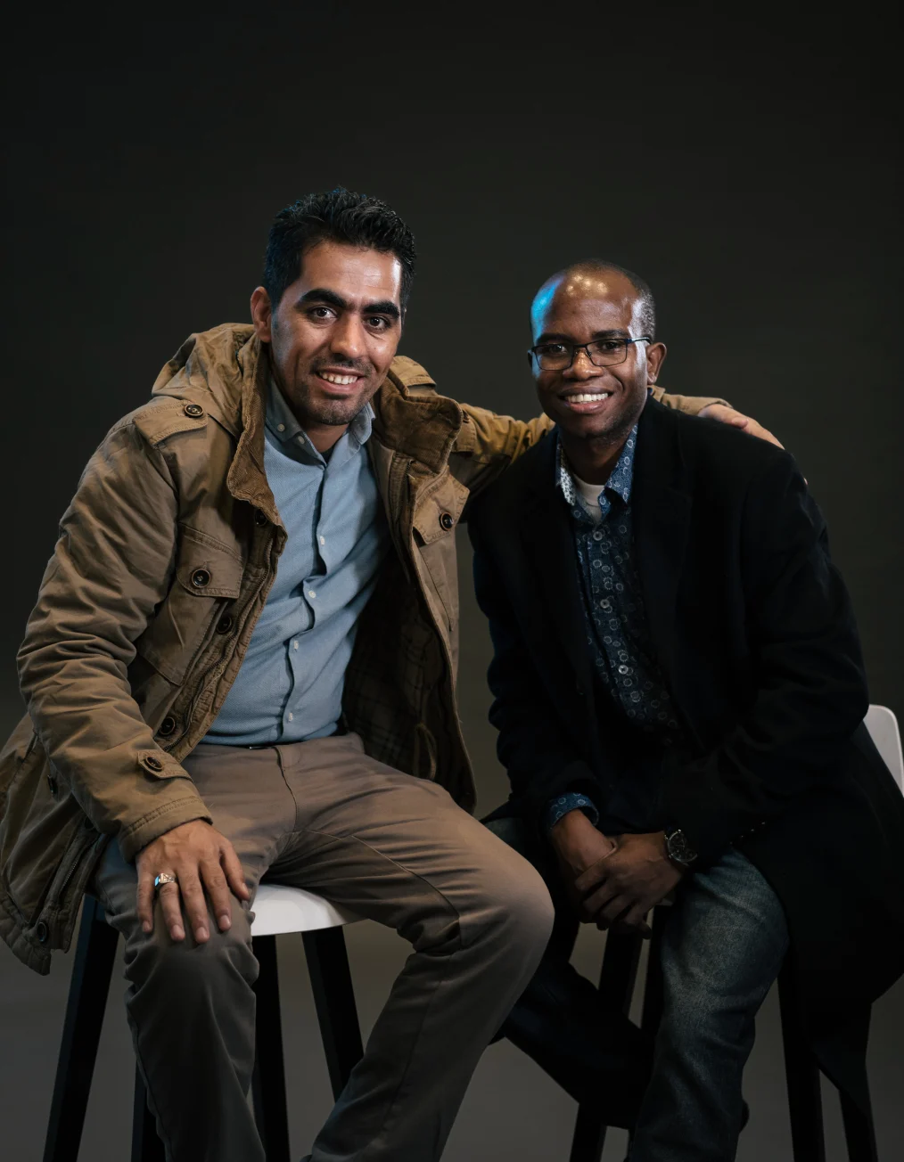 This picture shows a man with a medium skin tone and a man with a deep skin tone with dark hair 
sitting in white chairs smiling toward the camera.