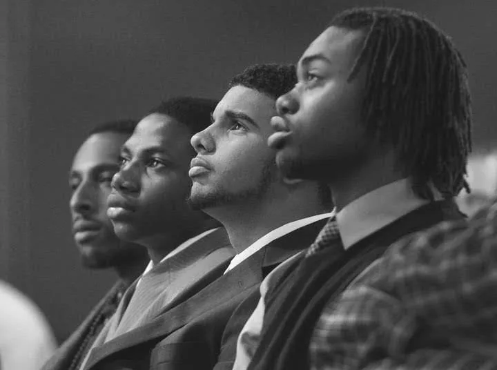 Four young men are pictured in black and white staring left out of frame.